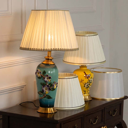 Beige pleated shade floral ceramic table lamp on wooden sideboard with light on for vintage decorative décor.