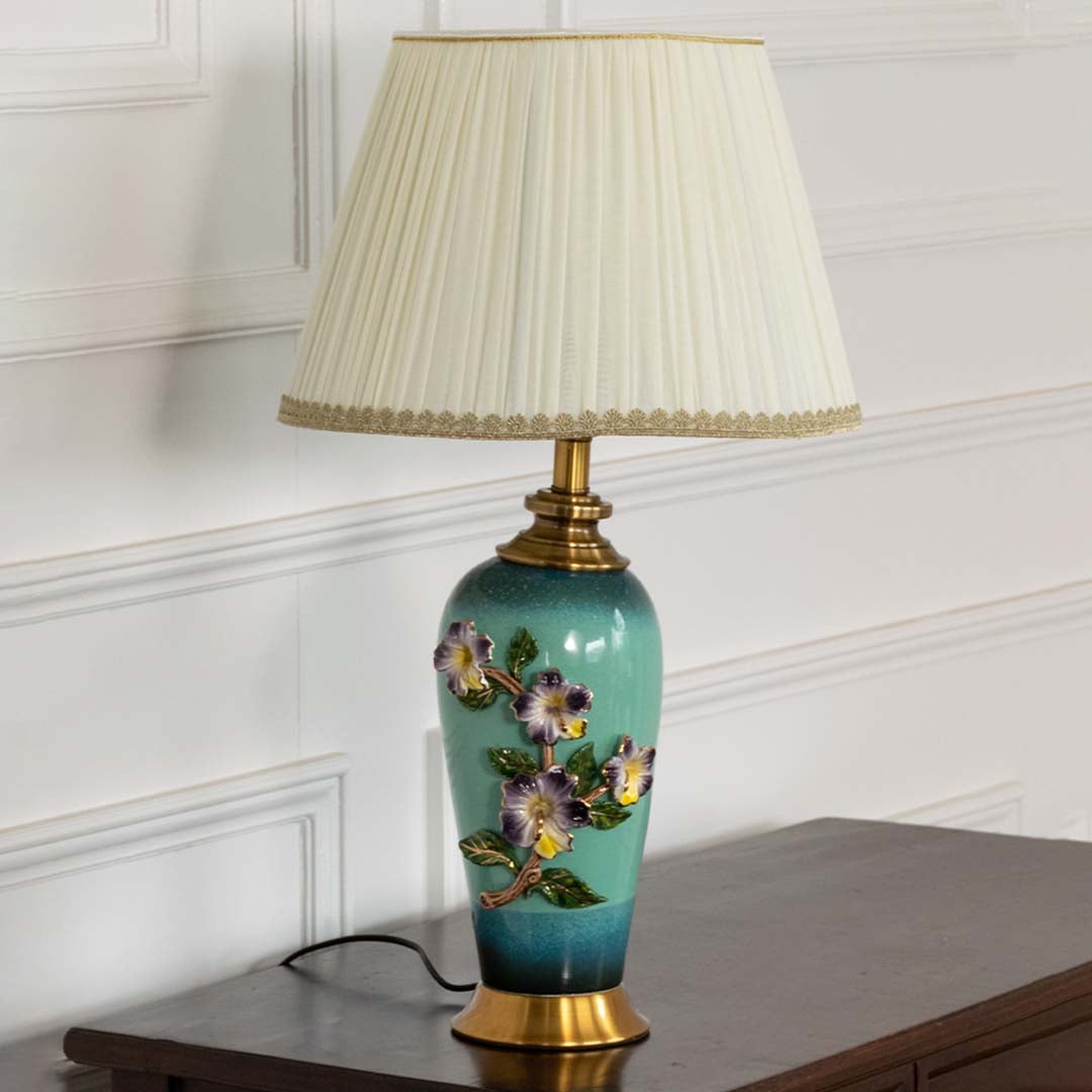 Pleated shade floral ceramic table lamp on wooden side table with light on for vintage decorative home décor.
