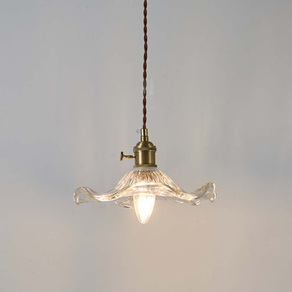 Clear wavy glass pendant light with brass holder glowing warmly on white background for decorative home lighting.