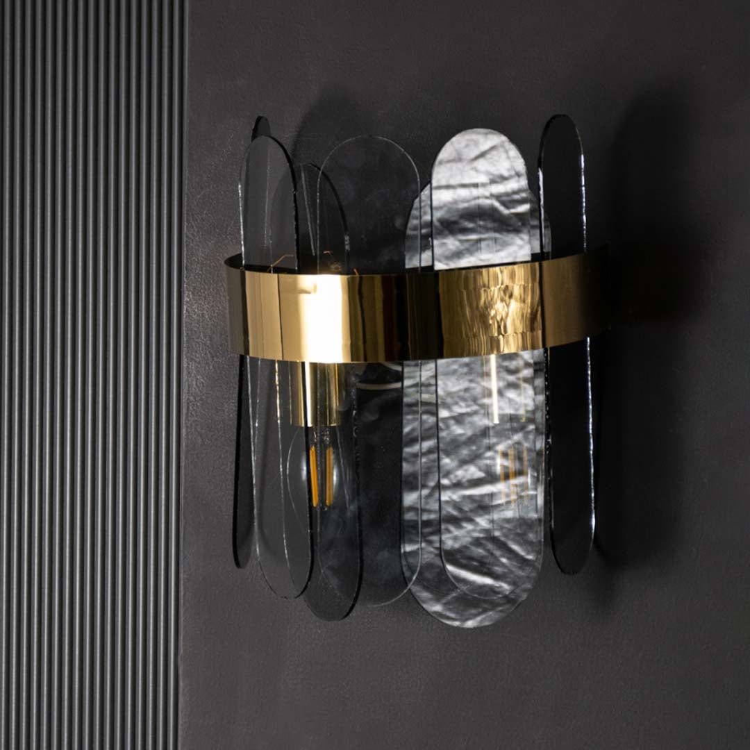 A metal and smoked glass wall light with bulbs on casting a warm glow on a dark black backdrop highlighting its modern layered glass panels.