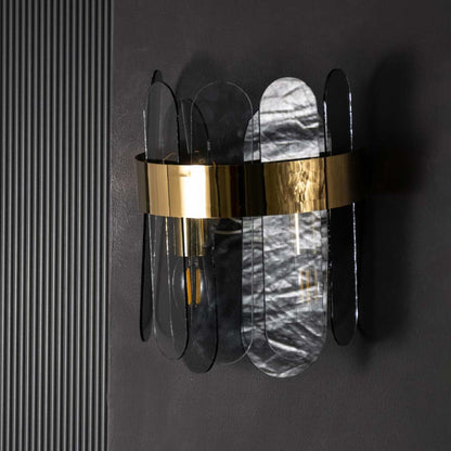 A metal and smoked glass wall light with bulbs on casting a warm glow on a dark black backdrop highlighting its modern layered glass panels.