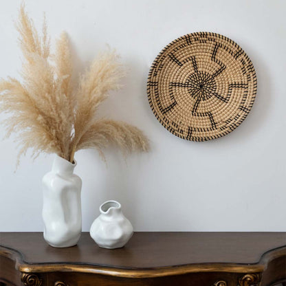 Handwoven wall basket decor in natural beige with black swirl design styled above console table with vases for boho wall.