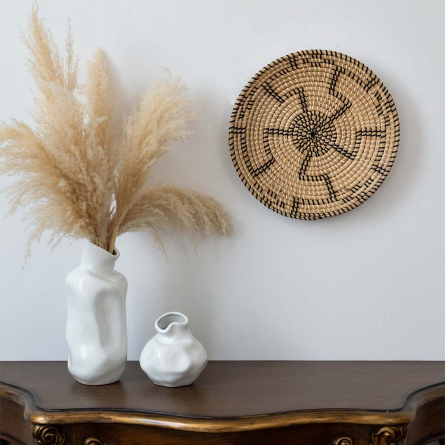 Handwoven wall basket decor in natural beige with black swirl design styled above console table with vases for boho wall.