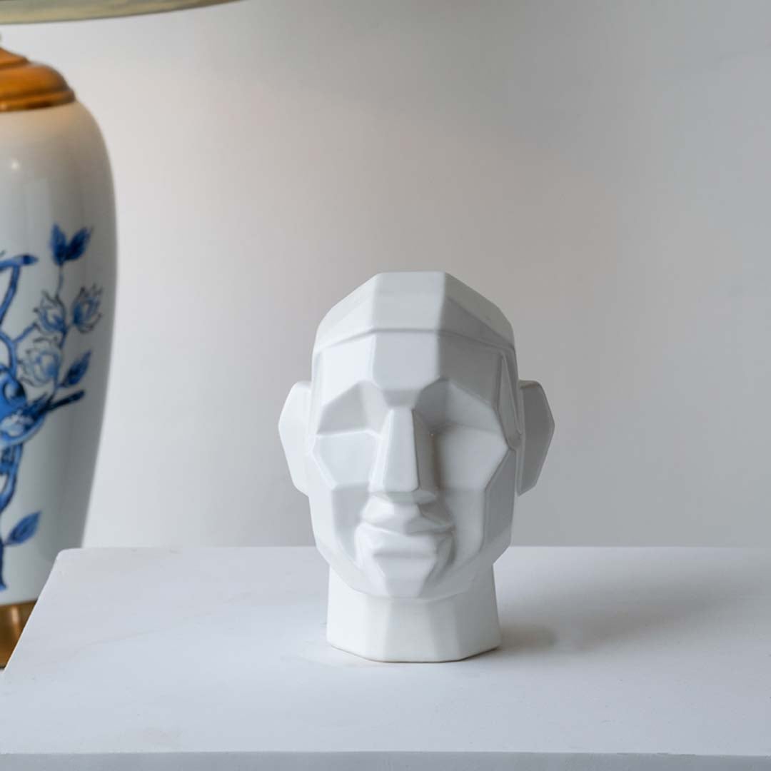 Modern geometric face sculpture displayed on a minimal white platform with soft natural light creating subtle shadows and depth.