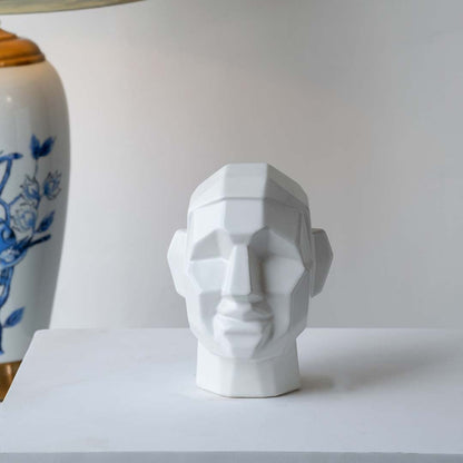Modern geometric face sculpture displayed on a minimal white platform with soft natural light creating subtle shadows and depth.