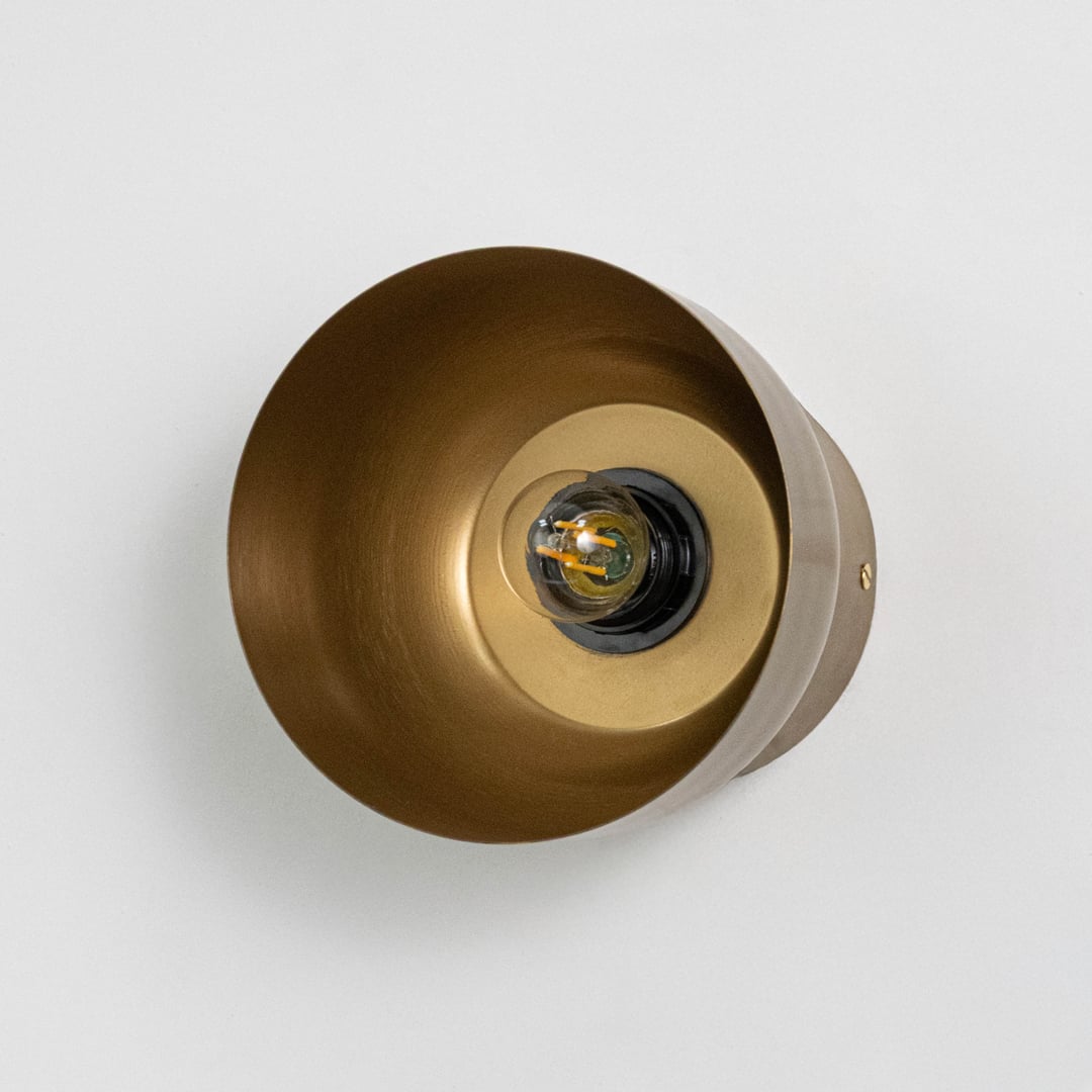 Brass cylindrical wall lamp with filament bulb on light background shown front facing.