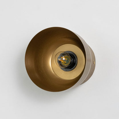Brass cylindrical wall lamp with filament bulb on light background shown front facing.
