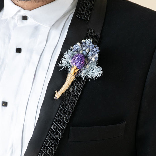 Detailed close up view of an artisanal blue and purple flower boutonniere pinned to a luxury black suit jacket lapel panel.
