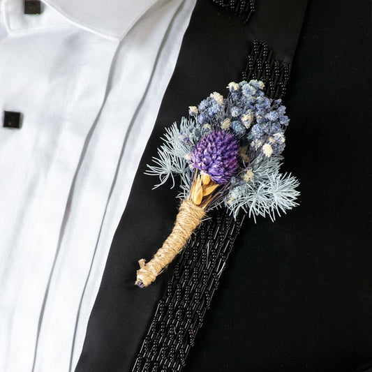 Closeup view of dried flower boutonniere with purple and blue blooms tied in jute placed on black suit for wedding attire.