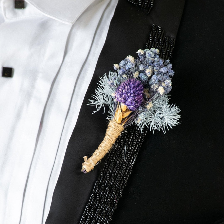 Closeup view of dried flower boutonniere with purple and blue blooms tied in jute placed on black suit for wedding attire.