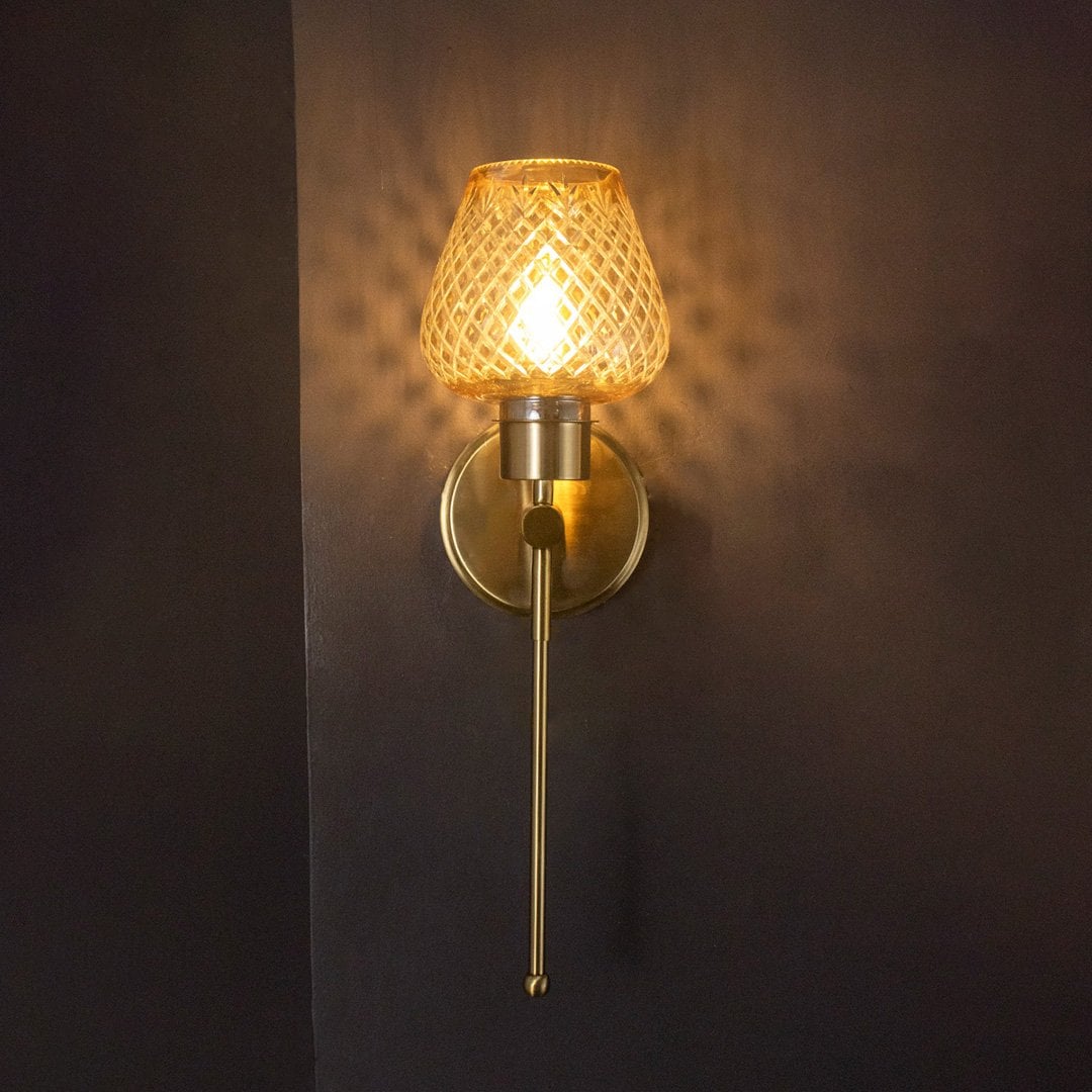 Glowing amber glass wall light with gold metal finish turned on suspended against a dark black background look.