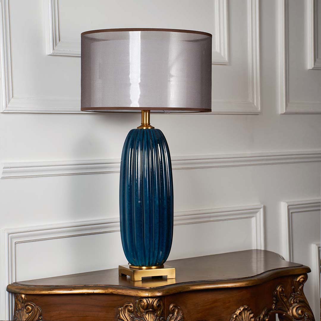 Tall blue ribbed ceramic lamp with gold base and grey drum shade shown unlit on a white background highlighting its modern bold shape.