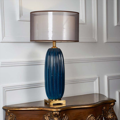 Blue ceramic ribbed table lamp with gold base and glowing grey shade displayed on a clean white background for premium decor styling.