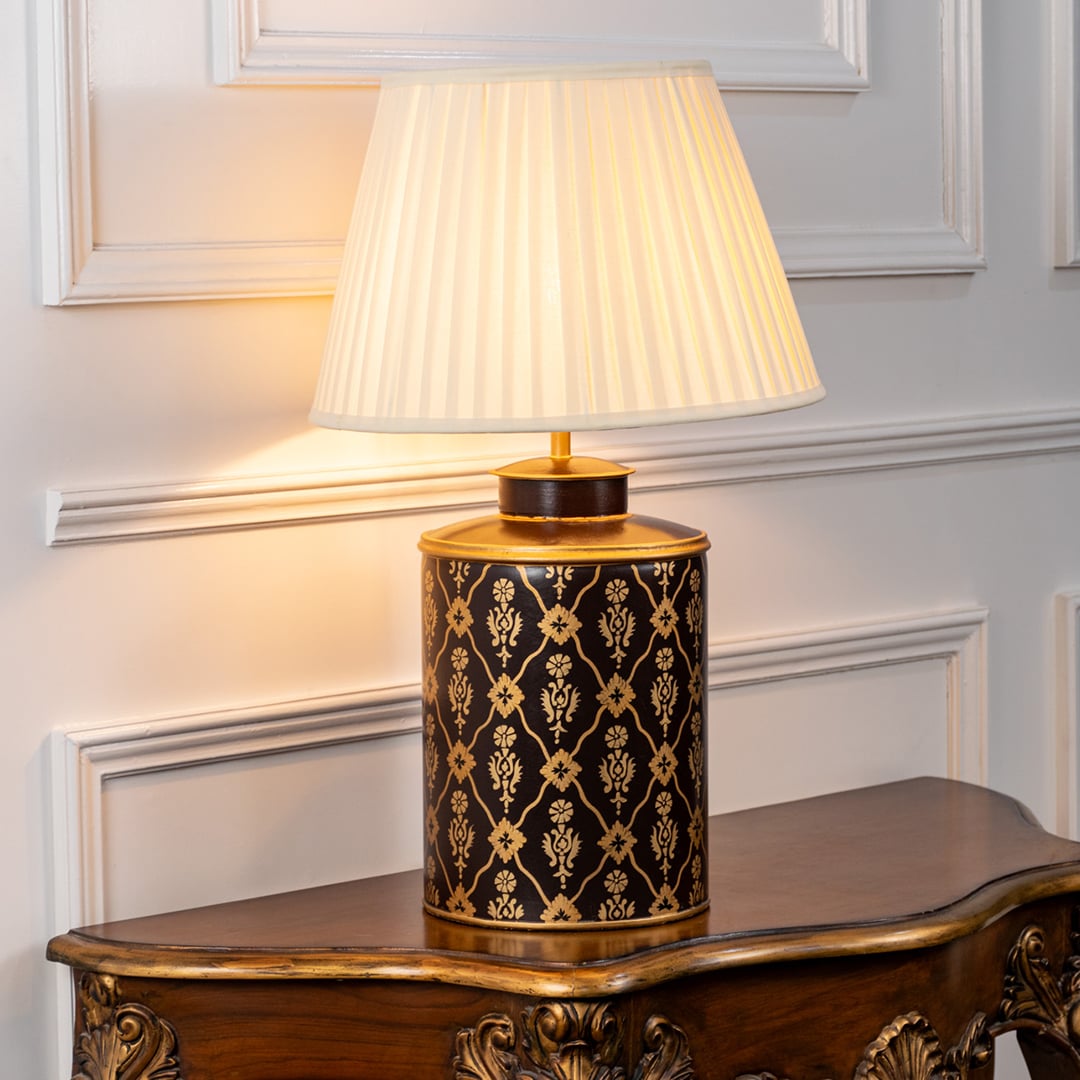 Table lamp on white background highlighting black and gold patterned ceramic base finished with a pleated fabric lampshade.