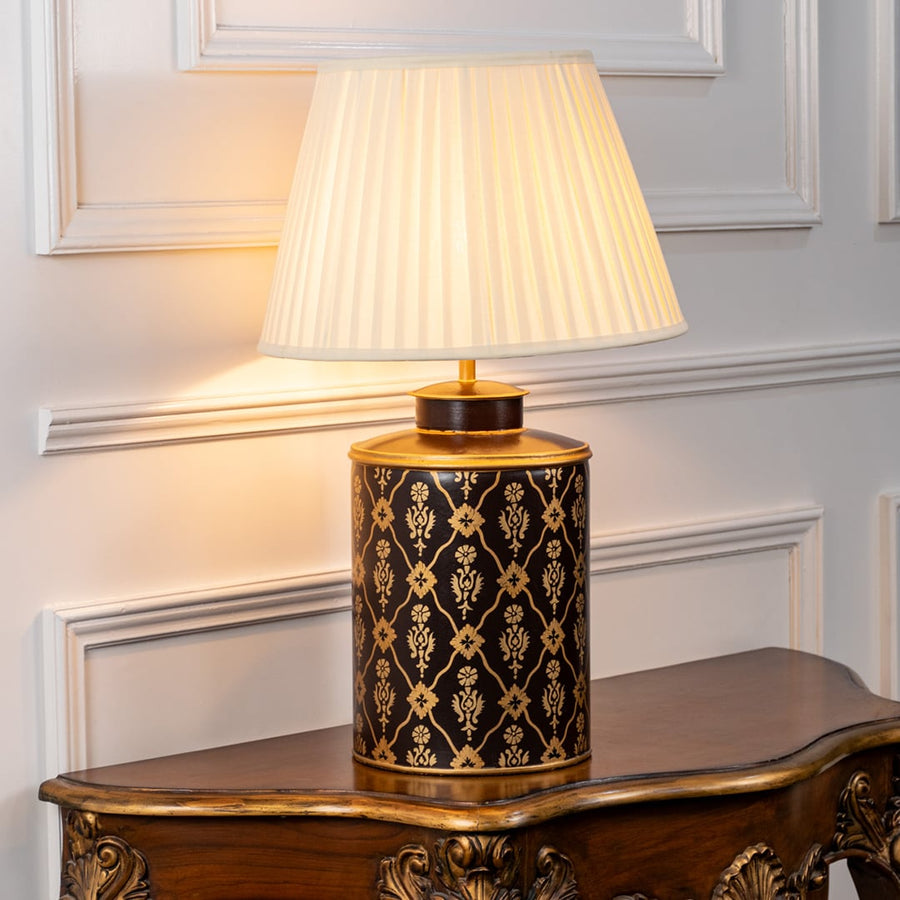 Table lamp on white background highlighting black and gold patterned ceramic base finished with a pleated fabric lampshade.