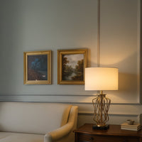 Gold metal table lamp with an artistic twisted base and warm white drum shade placed on a wooden side table creating soft ambient lighting in an elegant living room.