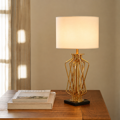 A modern table lamp with a glowing white drum shade and a gold geometric wireframe base on a black square pedestal, set on a wooden table next to stacked books.