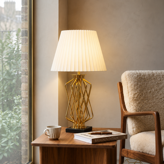 A modern table lamp with a pleated white shade and a geometric gold base sits on a wooden side table in a cozy room setting next to a window and a textured armchair.