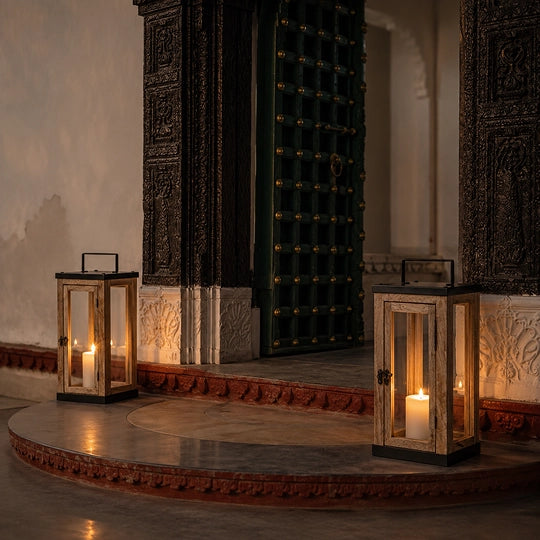 Two rustic wooden lanterns with black metal frames and lit candles, placed on a stone platform in front of an ornate dark green door with brass studs.