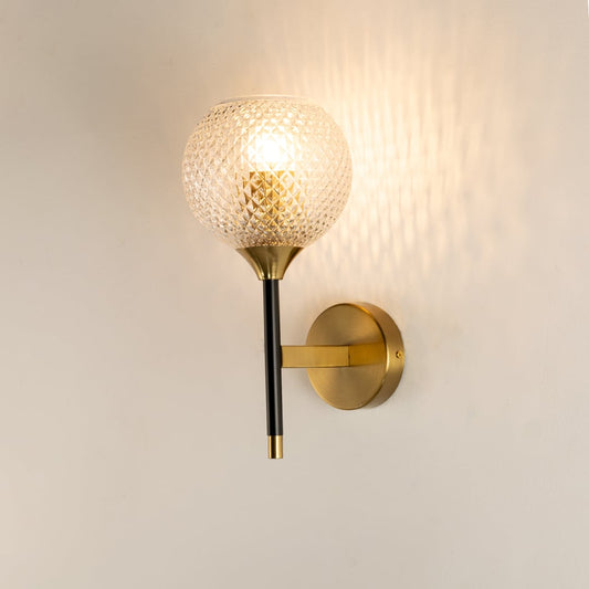 A gold and black wall light with a textured diamond cut glass globe shown with lights on against a white background highlighting its warm ambient glow.