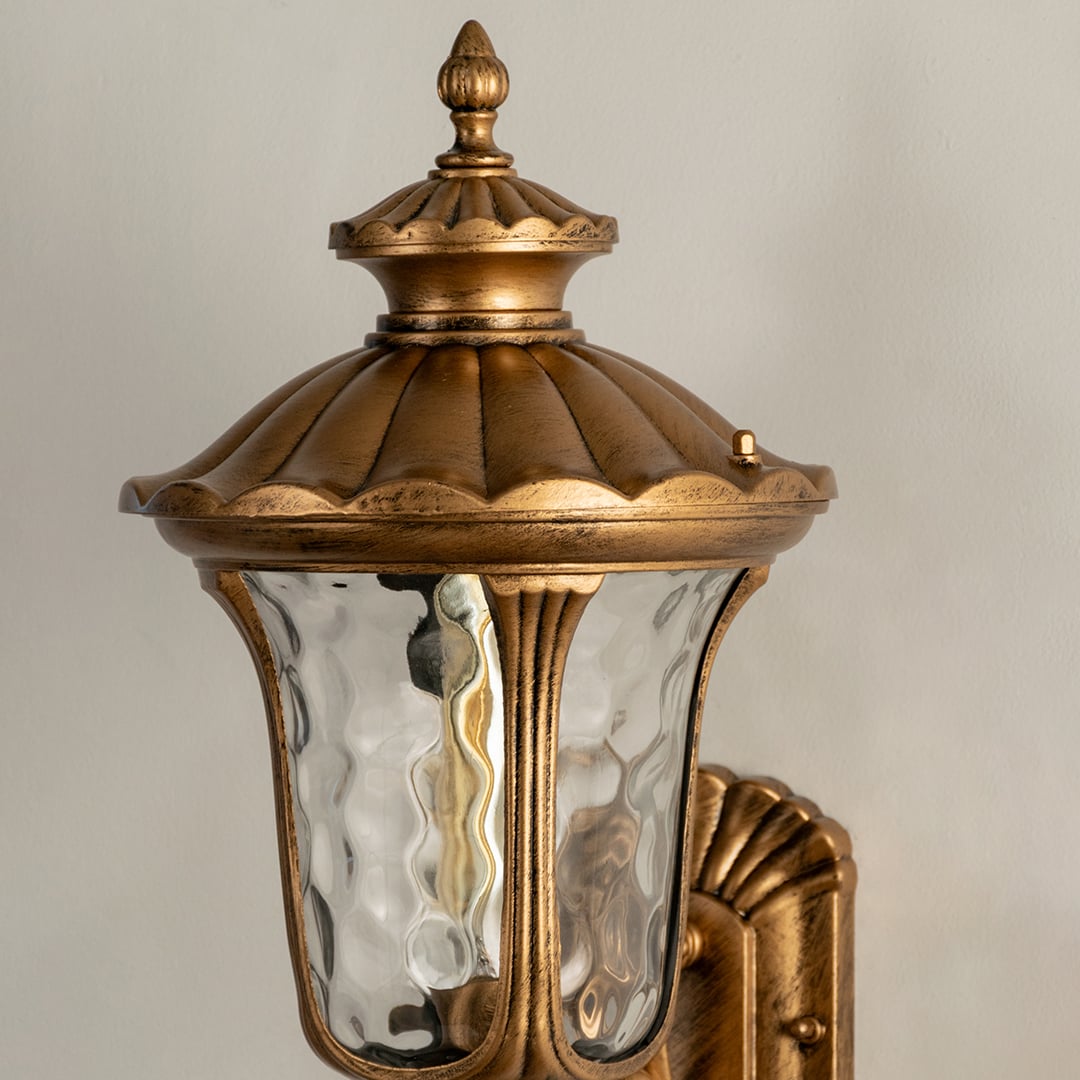 A closeup view of the gold outdoor lantern showing the warm glow through textured glass and highlighting the ornate carved details on the metal frame and roof.
