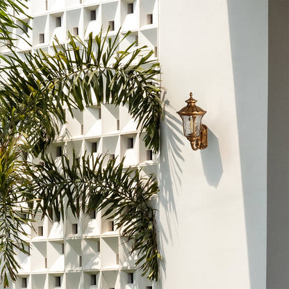 A classic gold outdoor wall lantern with the light off mounted on a bright white wall surrounded by palm leaves showcasing its ornate carved frame and elegant traditional style.