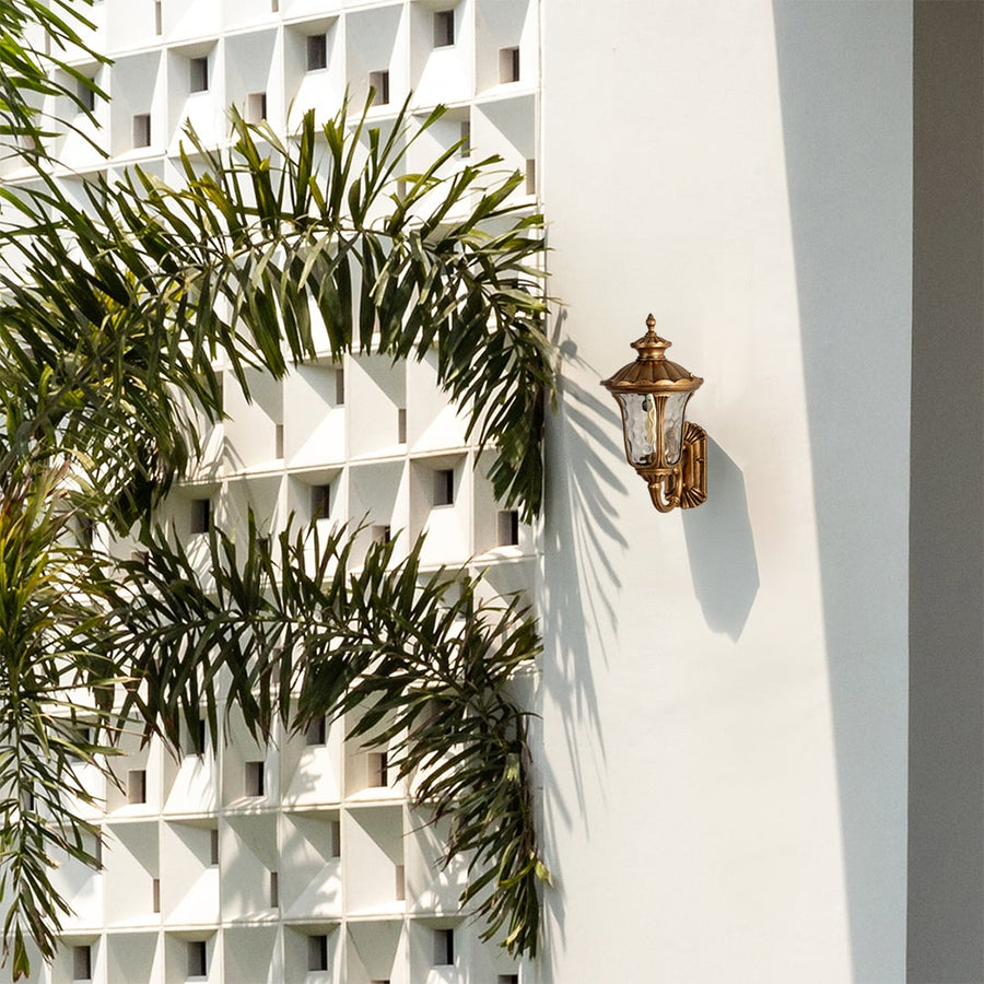 A classic gold outdoor wall lantern with the light off mounted on a bright white wall surrounded by palm leaves showcasing its ornate carved frame and elegant traditional style.