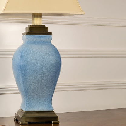 Closeup of cream shade blue ceramic table lamp on carved console table with light on and white panel wall background for classic elegant décor.