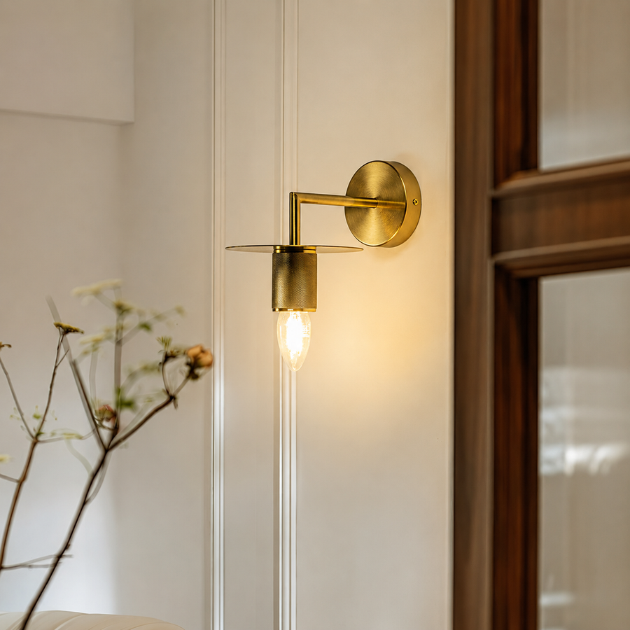 A modern wall-mounted sconce with a warm brass/gold finish, featuring a cylindrical shade and a glowing clear glass bulb, mounted on a white paneled wall next to a dark wood door frame.