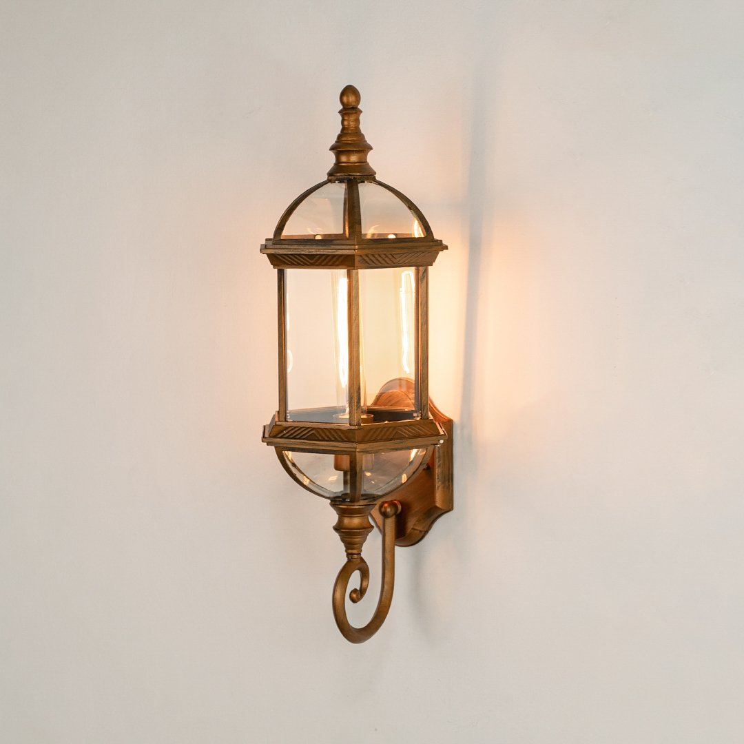 Antique brass metal outdoor wall light on white background with an elongated glass lantern and bottom scroll for decor.