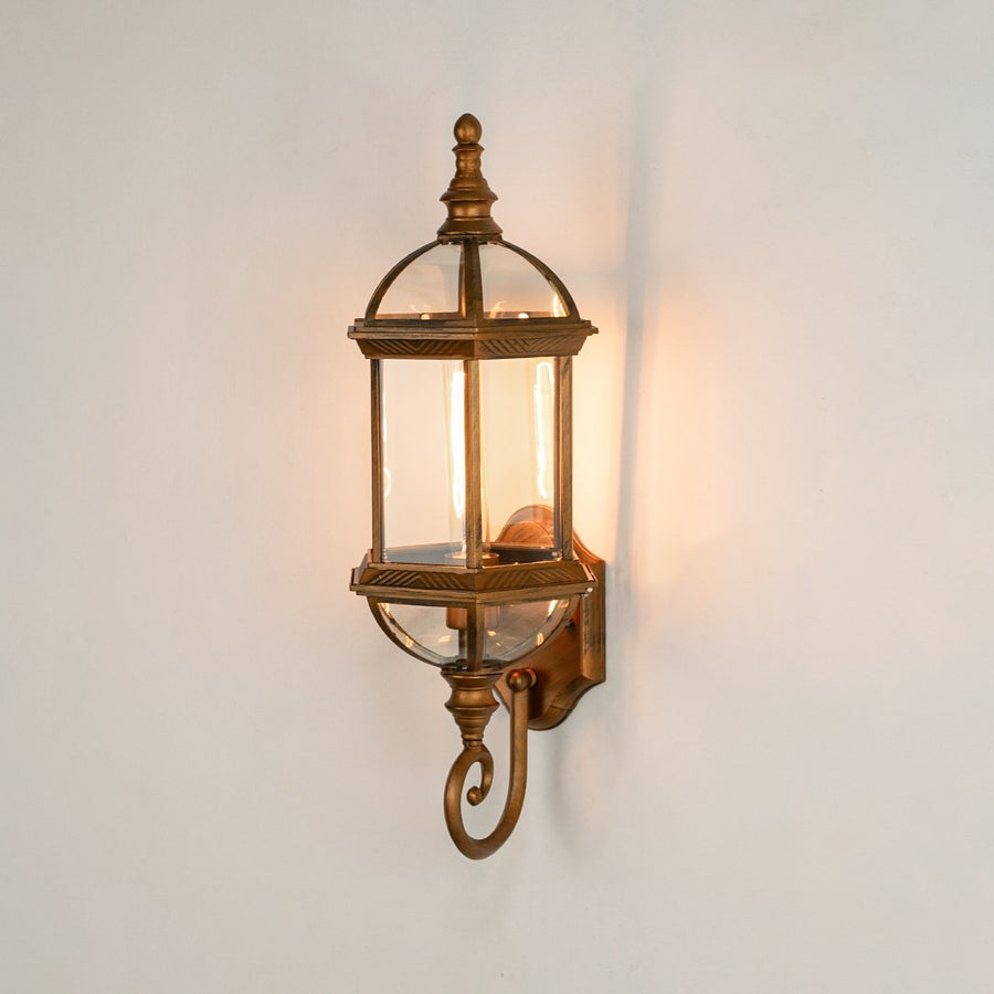 Antique brass metal outdoor wall light on white background with an elongated glass lantern and bottom scroll for decor.