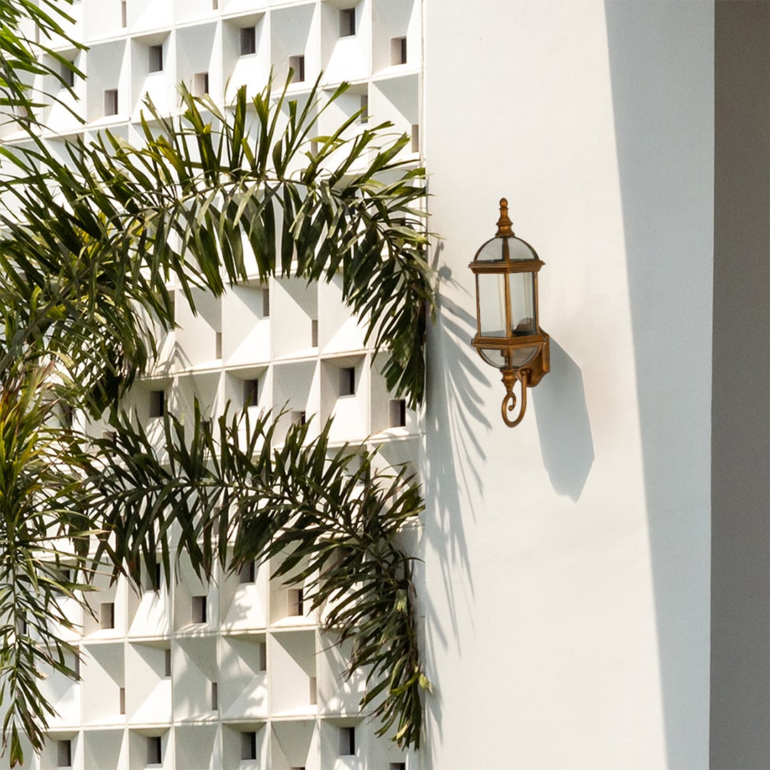 Classic bronze wall light fixture with glass panels off mounted on a white geometric patterned wall beside green palm trees.