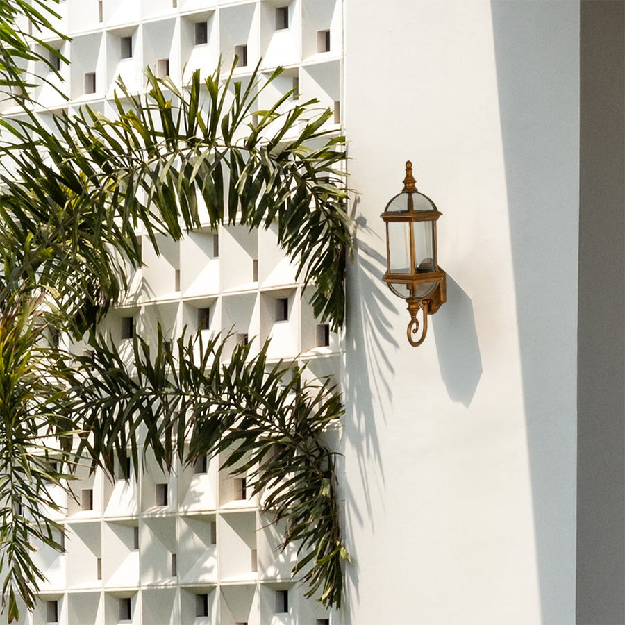 Classic bronze wall light fixture with glass panels off mounted on a white geometric patterned wall beside green palm trees.
