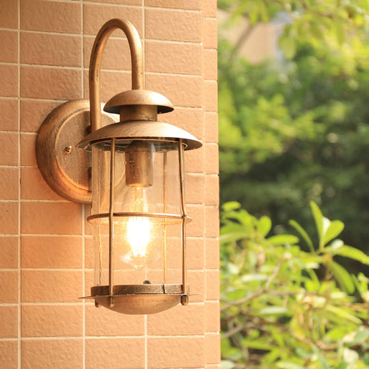 Industrial brass gooseneck outdoor wall sconce lifestyle image with clear glass cage lantern mounted on tan brick patio wall.