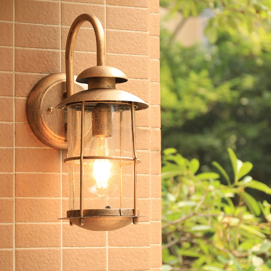 Industrial brass gooseneck outdoor wall sconce lifestyle image with clear glass cage lantern mounted on tan brick patio wall.