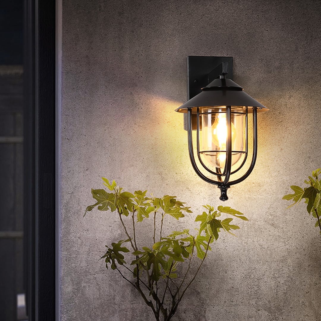 A warm outdoor scene featuring a black metal wall light glowing softly against a textured wall creating a cozy evening ambiance with plants adding natural charm.