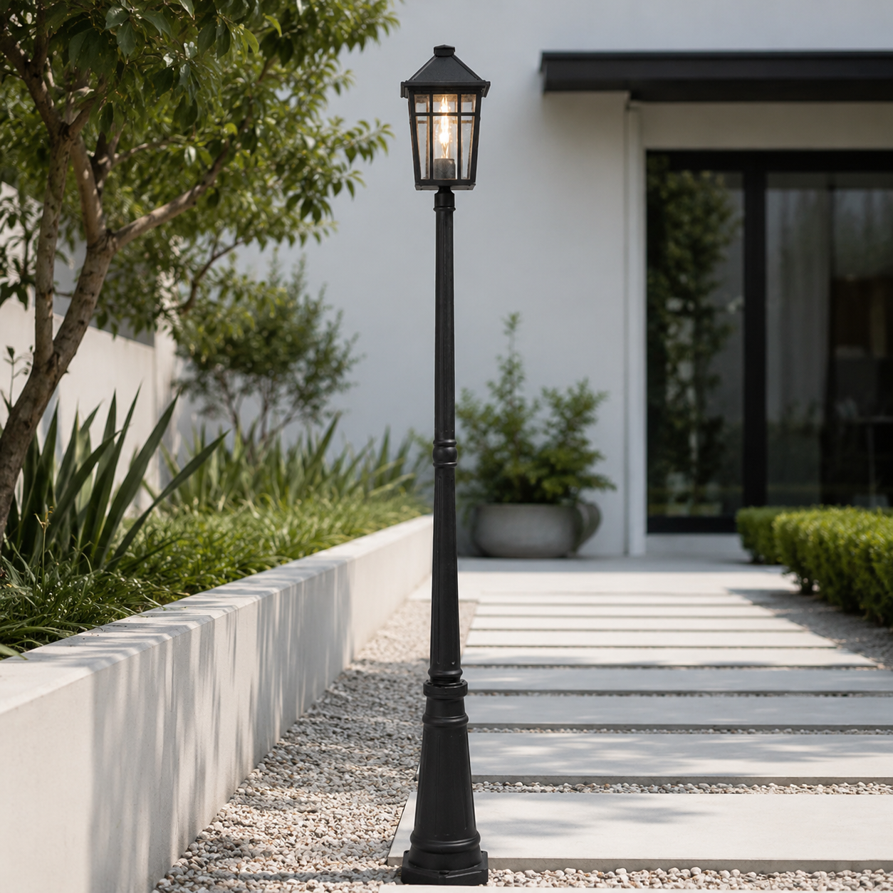 A black outdoor landscape light post with a lantern-style top, standing on a gravel path beside a modern white walkway and landscaped garden.