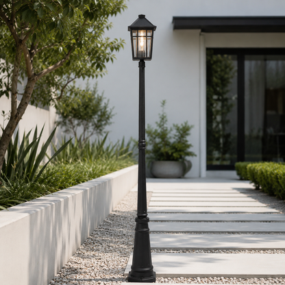 A black outdoor landscape light post with a lantern-style top, standing on a gravel path beside a modern white walkway and landscaped garden.