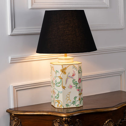 Table lamp with black fabric shade and cream ceramic base featuring delicate hand painted floral artwork displayed on a wooden table against a white background.