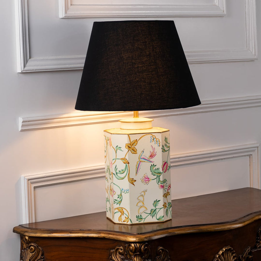 Table lamp with black fabric shade and cream ceramic base featuring delicate hand painted floral artwork displayed on a wooden table against a white background.