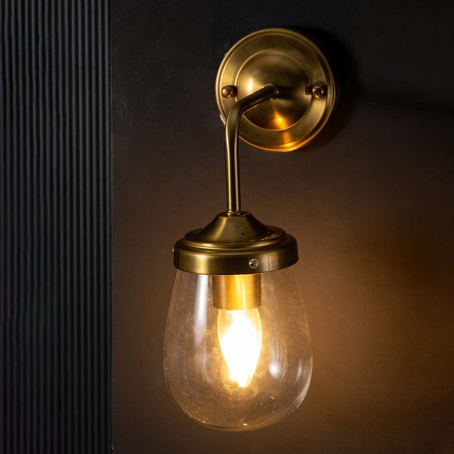 Gold wall sconce with a clear teardrop glass shade shown with the light on against a dark background producing a warm focused glow.