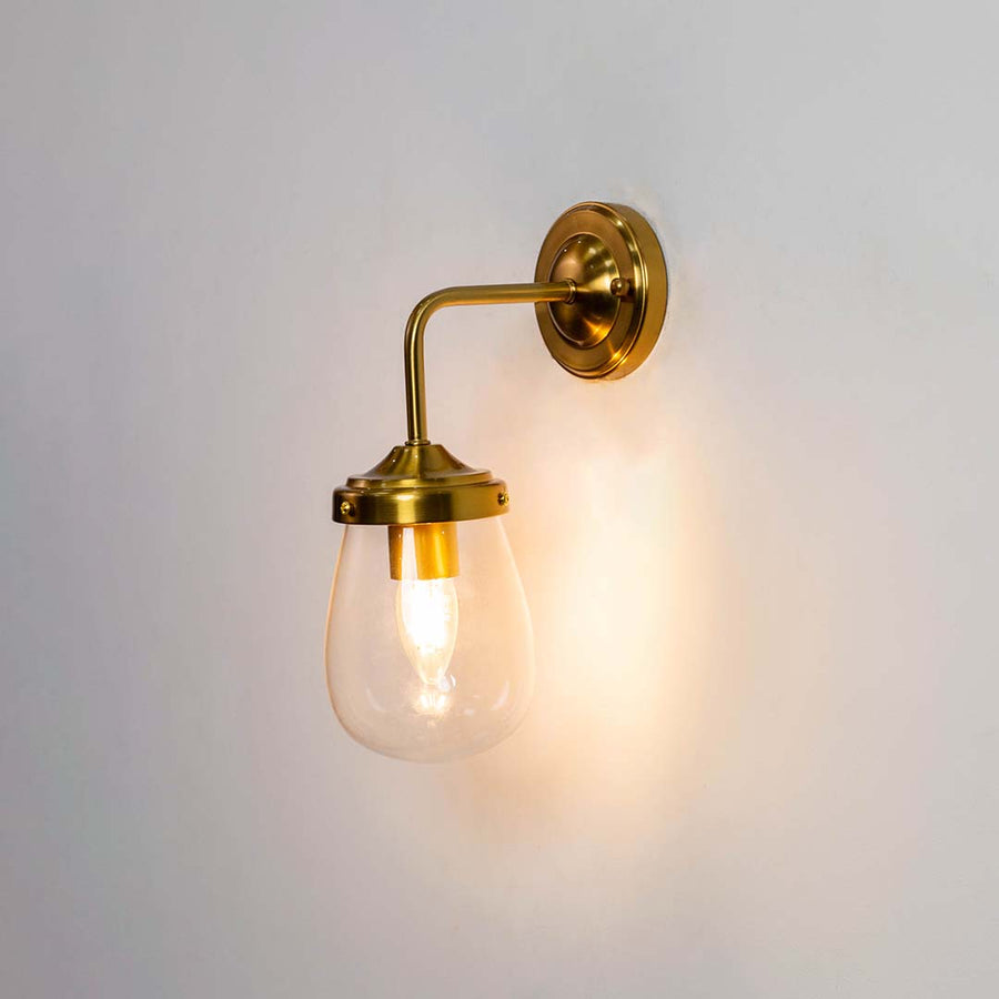 Gold wall sconce with teardrop glass shade shown with the light on against a clean white background creating a warm and inviting glow.