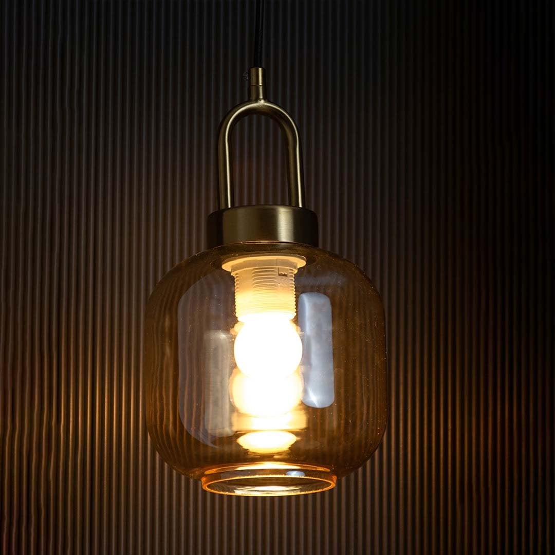 A gold and amber glass pendant light with lights on casting a warm glow on a black backdrop showcasing its tinted glass shade and modern brass finish.