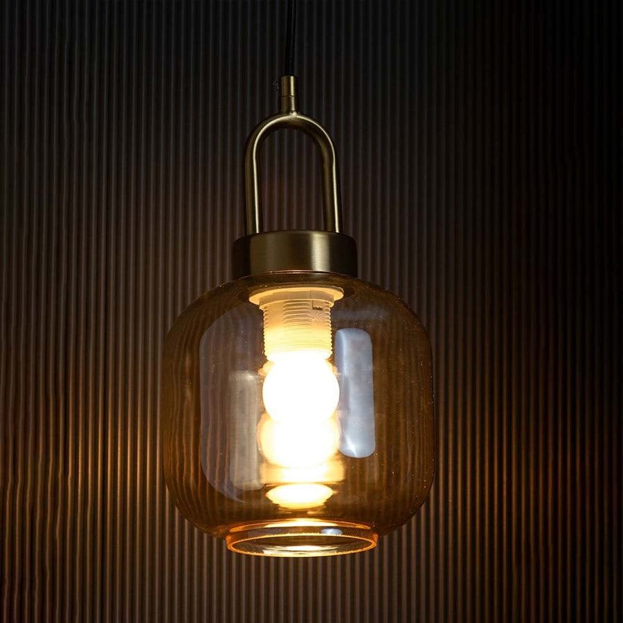 A gold and amber glass pendant light with lights on casting a warm glow on a black backdrop showcasing its tinted glass shade and modern brass finish.