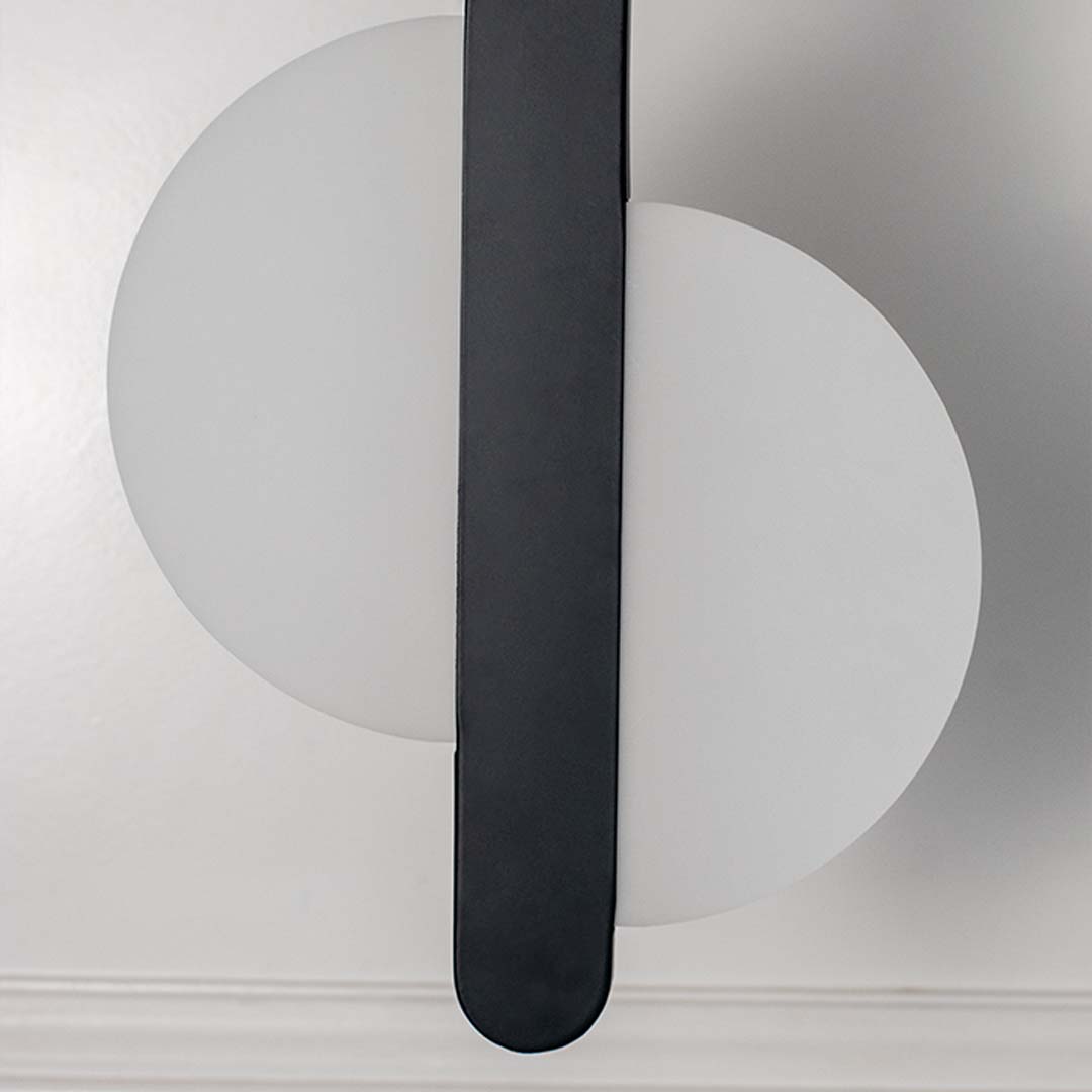Closeup modern wall light with white circular backplate and black vertical bar placed against neutral wall background showcasing minimal lighting decor styling.