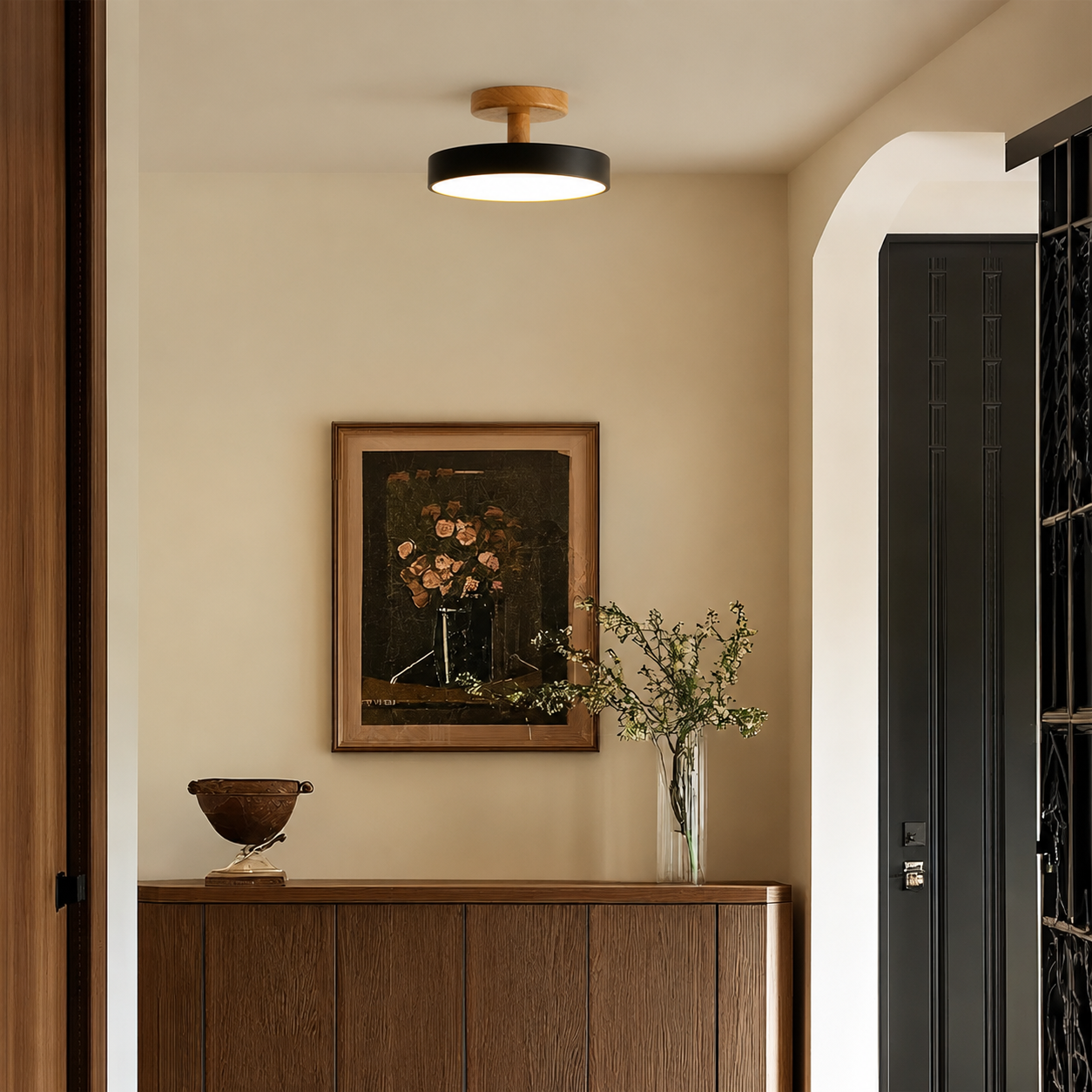 A modern ceiling light fixture with a black circular shade and a light wood top, mounted on a beige wall in a styled interior hallway above a wooden console table.
