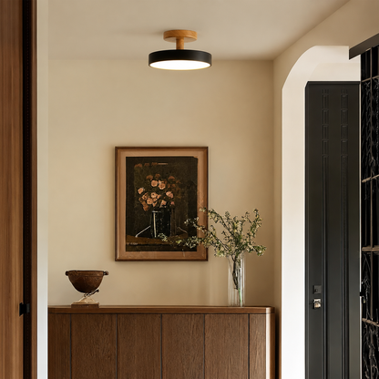 A modern ceiling light fixture with a black circular shade and a light wood top, mounted on a beige wall in a styled interior hallway above a wooden console table.