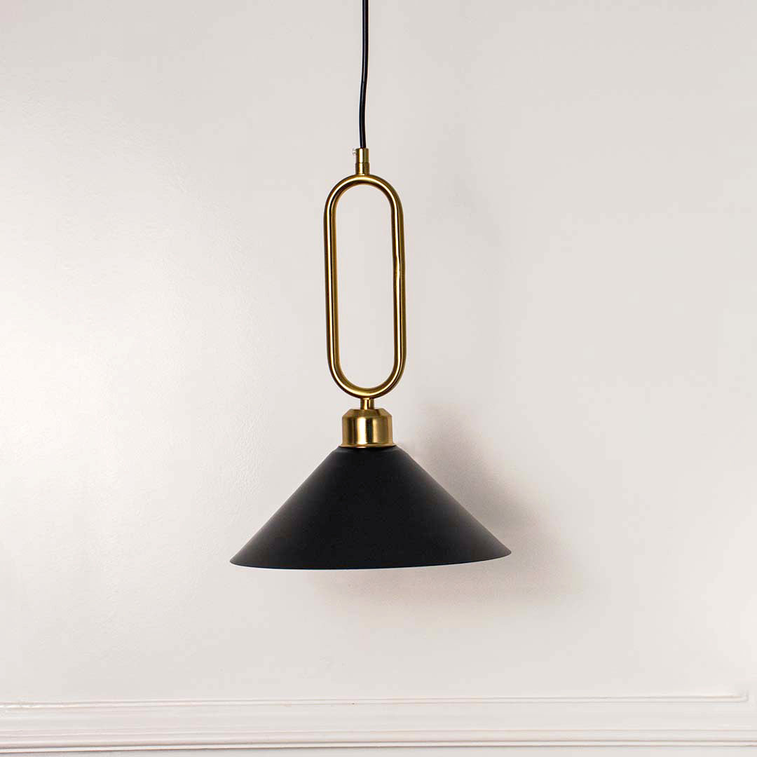Modern black metal pendant light with gold loop handle and cone shade hanging against soft beige wall background for elegant interior decor lighting.