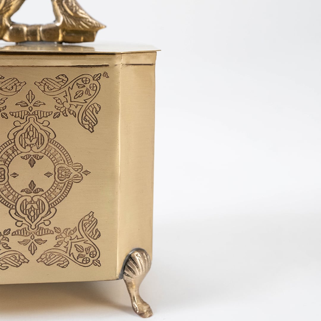 Main image of decorative gold metal box with bird handle and engraved floral sides placed on white background.