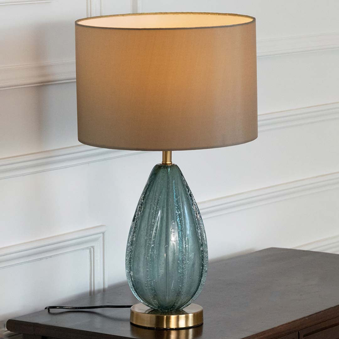 Teal glass table lamp with fabric shade displayed on a wooden console table creating elegant ambient lighting indoors.